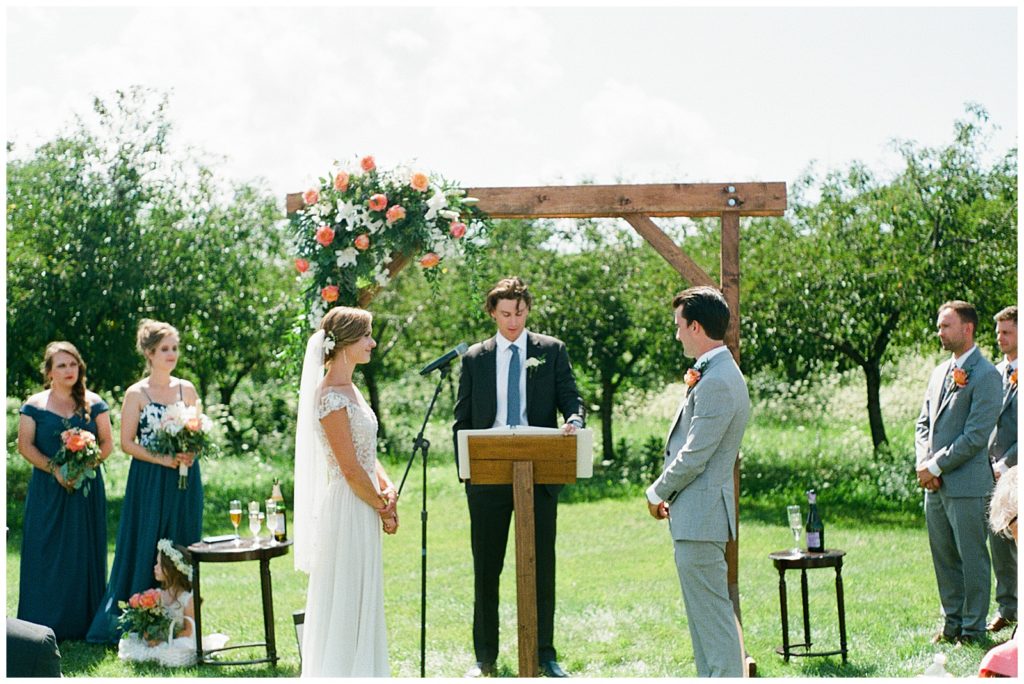 In wedding photography on film, a couple gets married under an arbor with pink and white flowers.