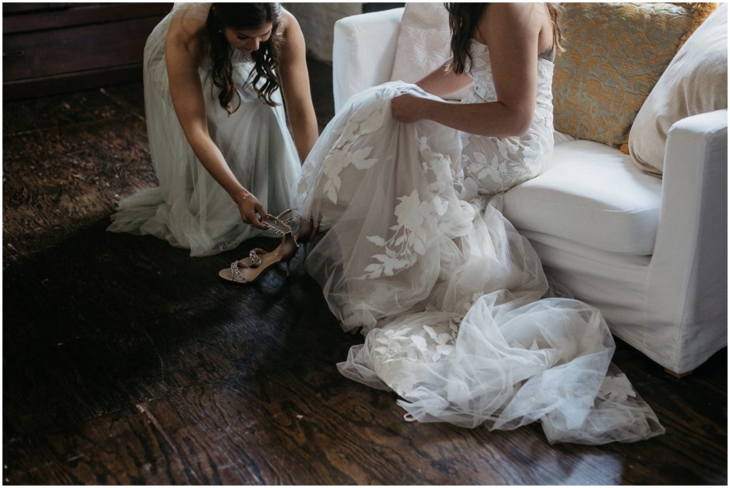 bridesmaid helps bride put on shoes for wedding on weekday