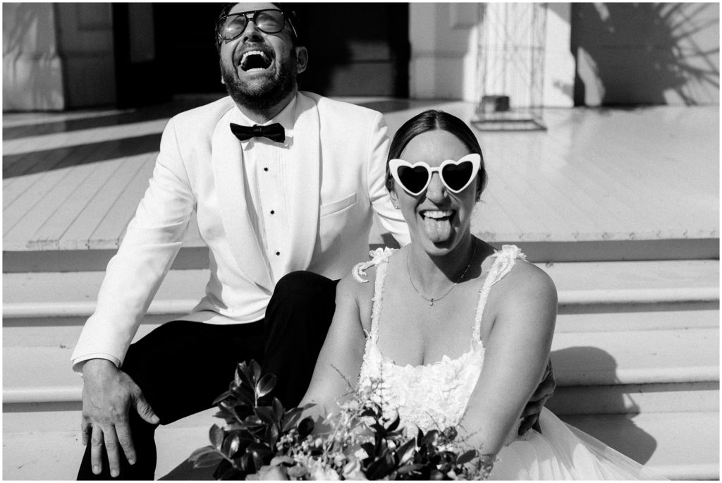 A bride and groom sit on the steps of Derbes Mansion wearing sunglasses and making funny faces.