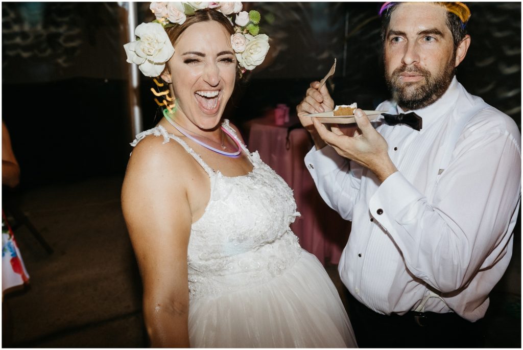 A bride laughs while a groom eats cake.