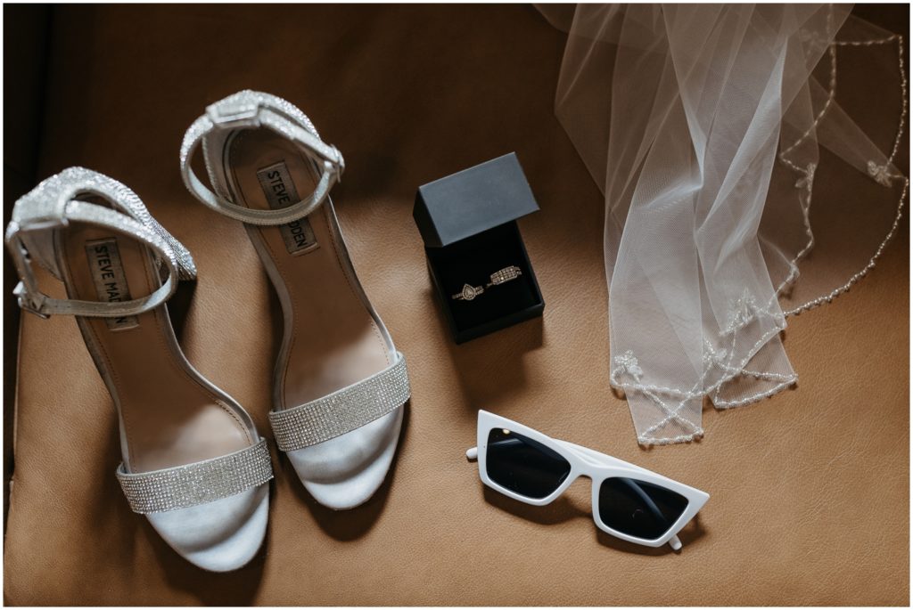 Shoes, wedding rings, and sunglasses sit on a tan floor.