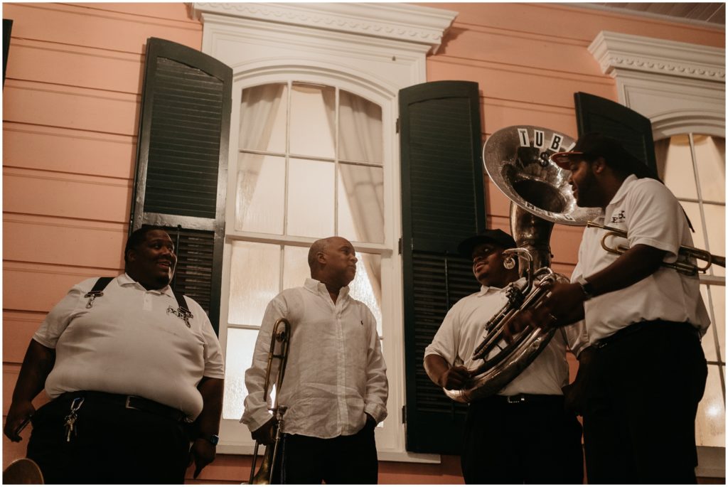 A brass band stands in front of a pink and green New Orleans house.