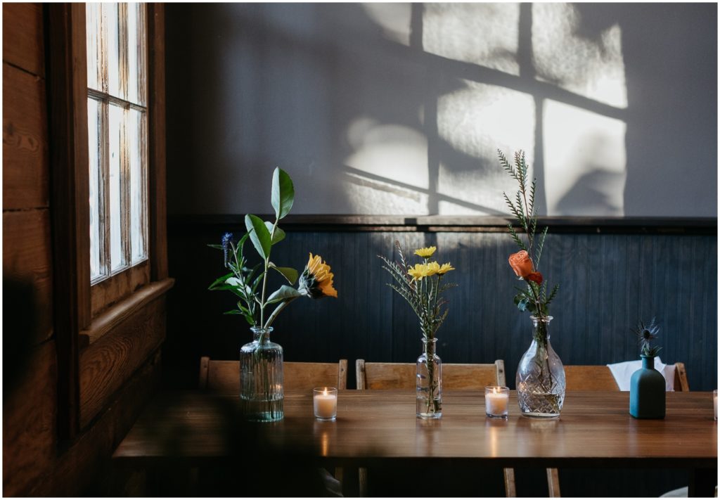 Flowers sit in small vases on a wooden table beside a window.