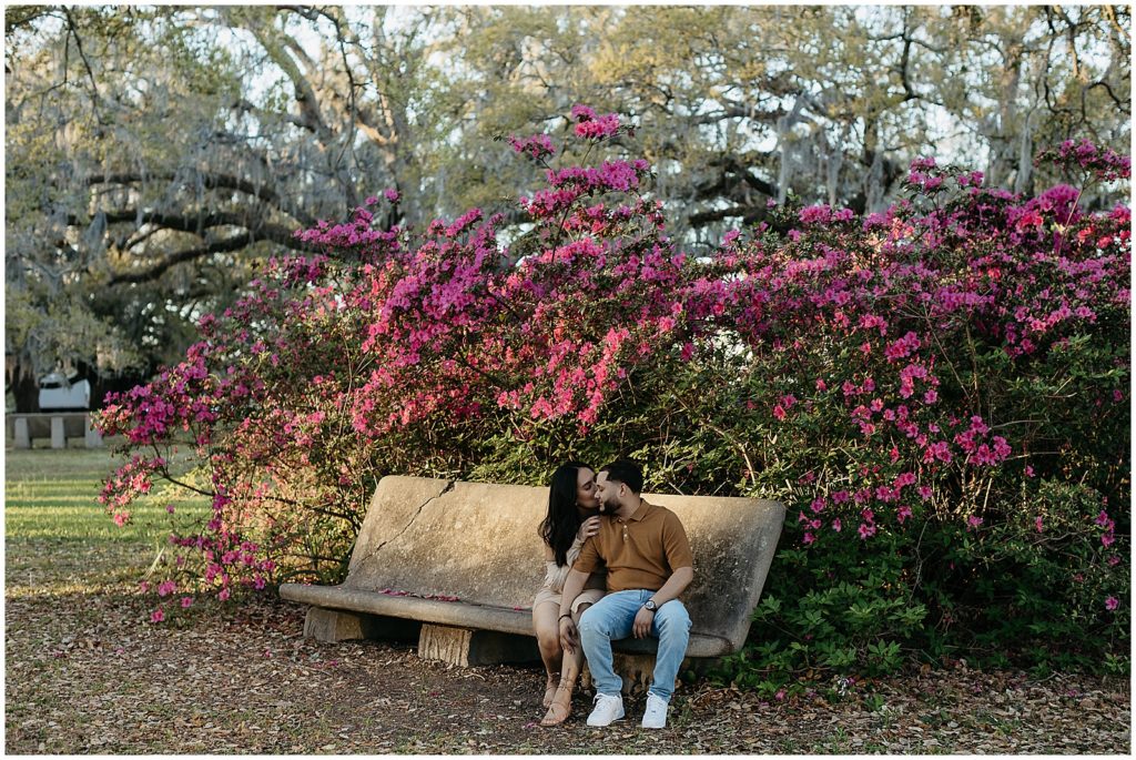 Pink flowers grow over a bench where a couple sits holding hands.
