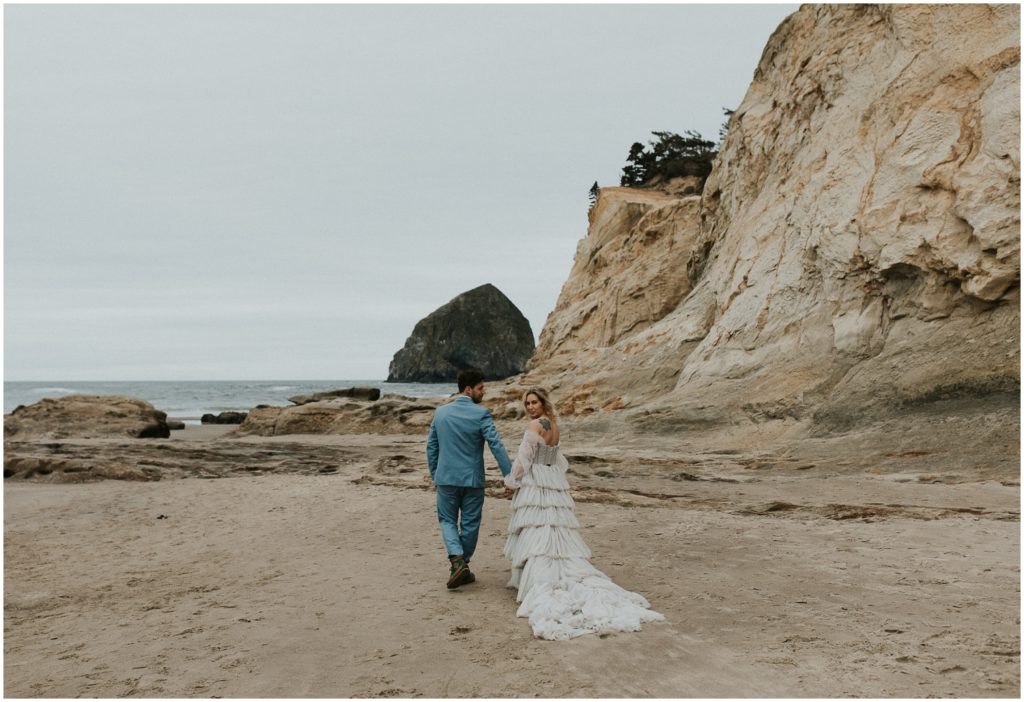 A bride holds her husband's hand and looks over her shoulder while they walk away from the camera.