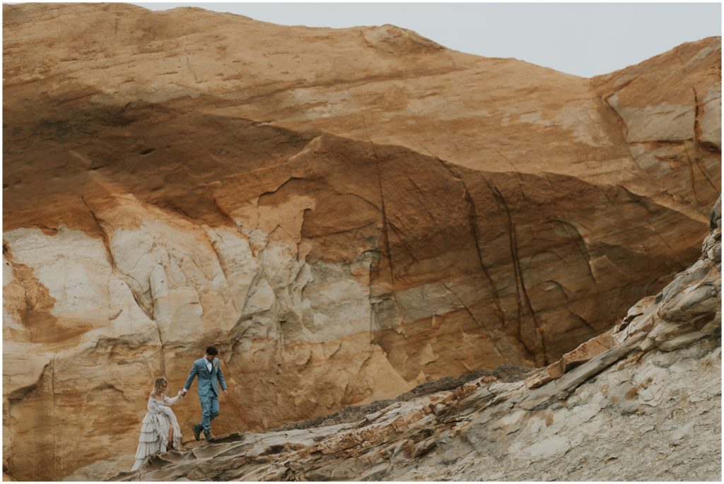 A couple in blue wedding attire hikes up an orange and brown sandstone cliff at Cape Kiwanda.