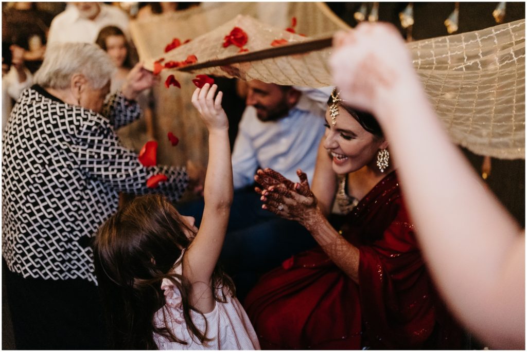 Wedding guests sprinkle flower petals on a veil over a seated bride and groom.