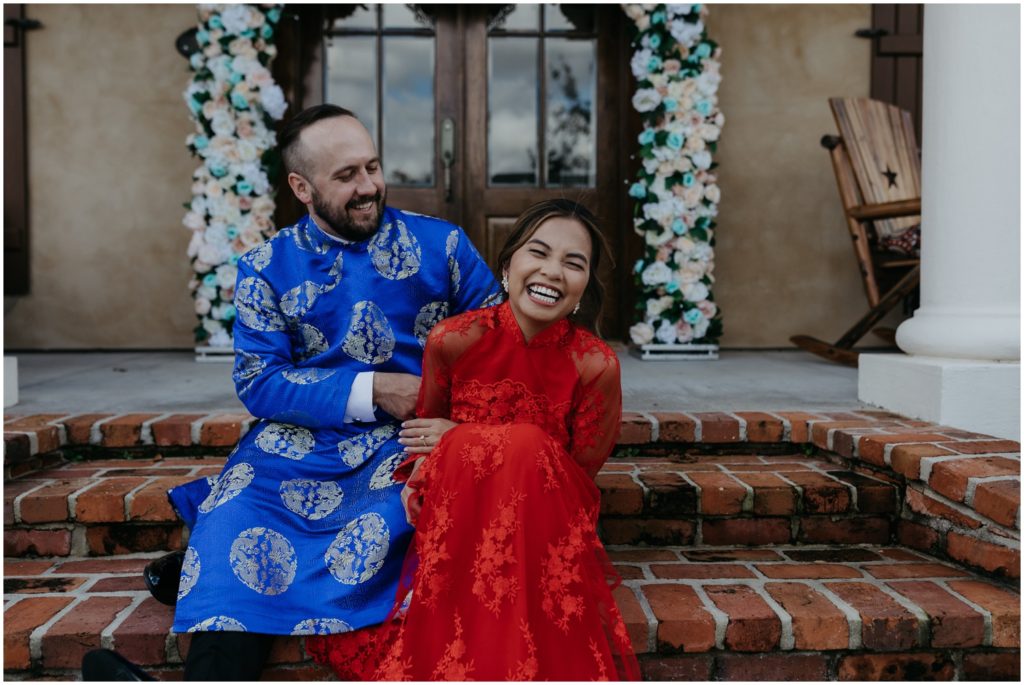 A bride and groom in Vietnamese wedding attire laugh on their porch steps.