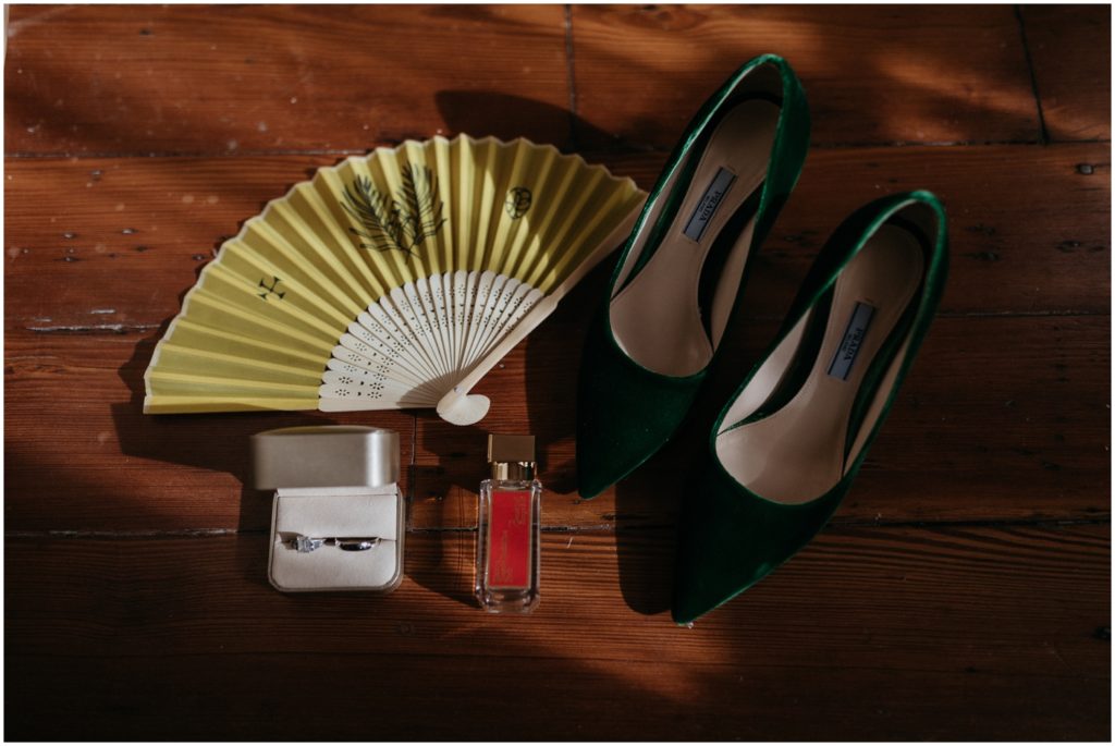 A pair of green wedding shoes sits beside a gold fan on the floor of a getting ready suite.