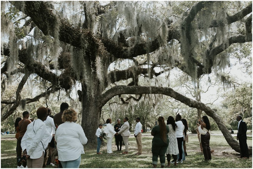 Two women have their elopement ceremony under an oak tree as family members look on.
