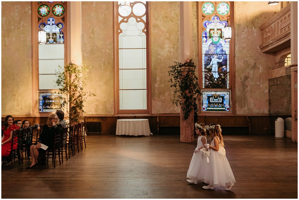Flower girls walk up the aisle with stained glass windows behind them.