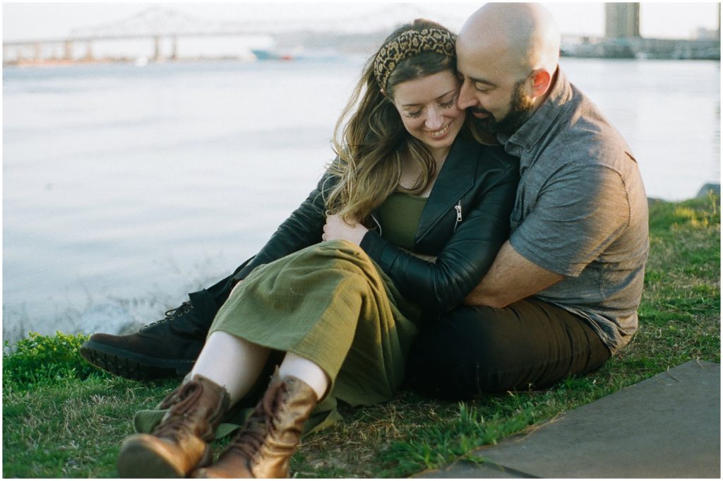 An engaged couple sits together on the levee in New Orleans with the bridge in the background.