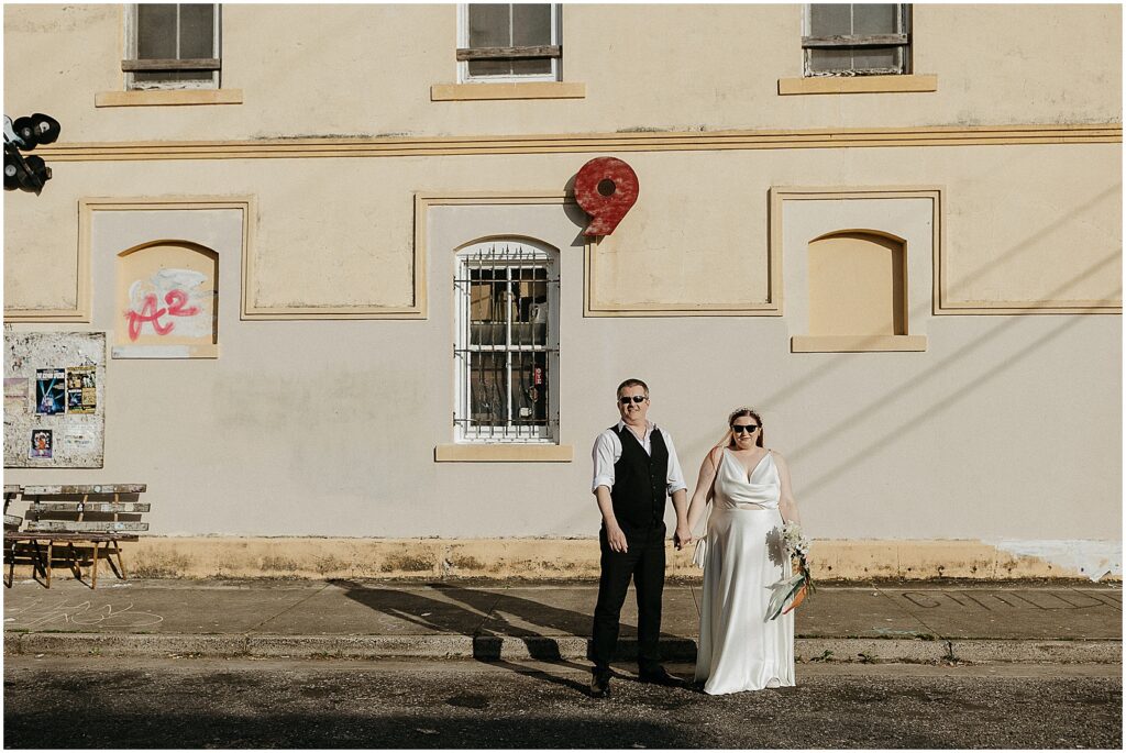 A bride and groom pose for a New Orleans wedding photographer in front of a yellow building.