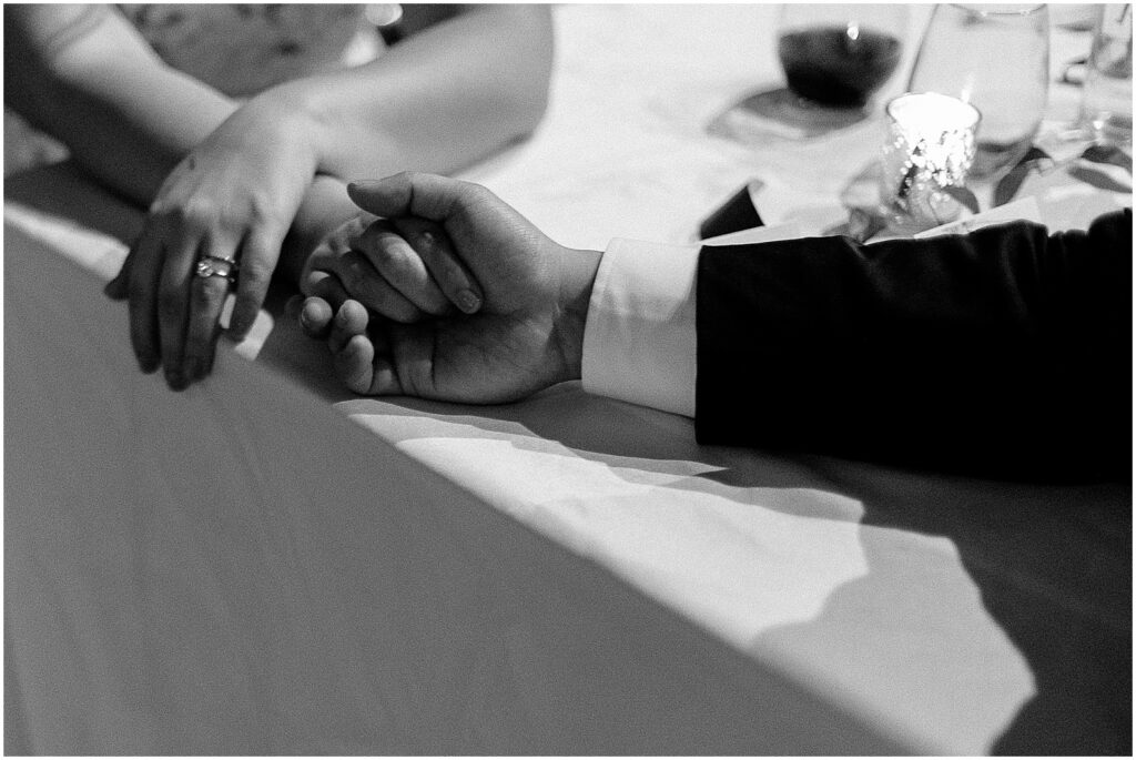 A bride and groom hold hands at their reception table.