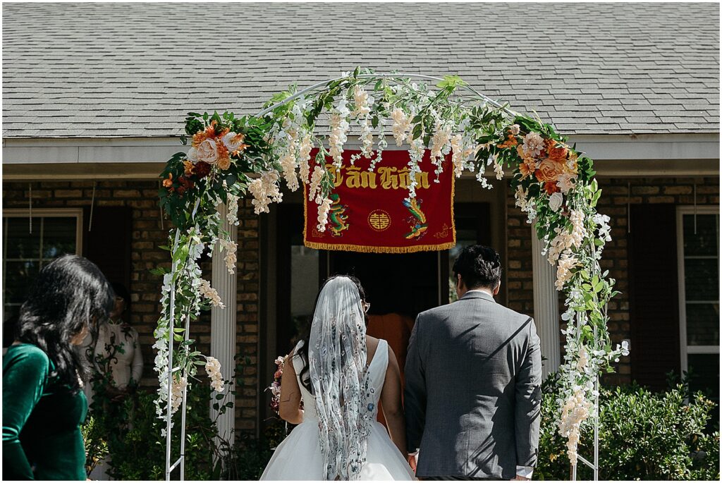 A bride and groom walk under a floral arch towards a Viet prayer ceremony before their New Orleans wedding.