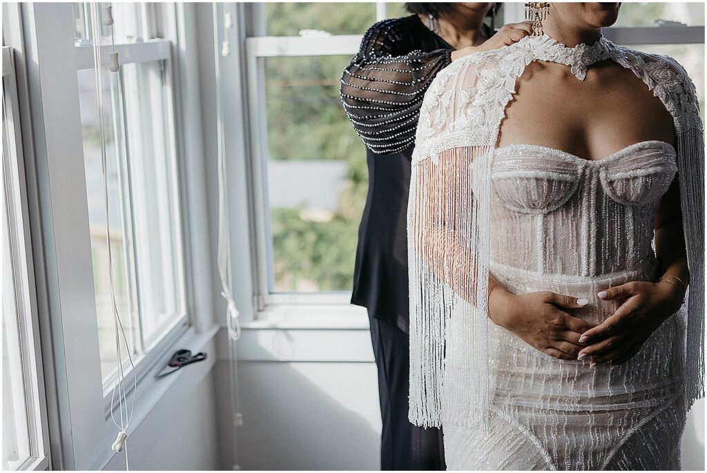 A family member fastens a bride's wedding cape beside a window.