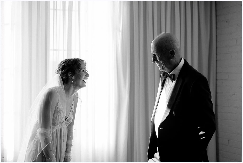 A bride and her father laugh beside a window.