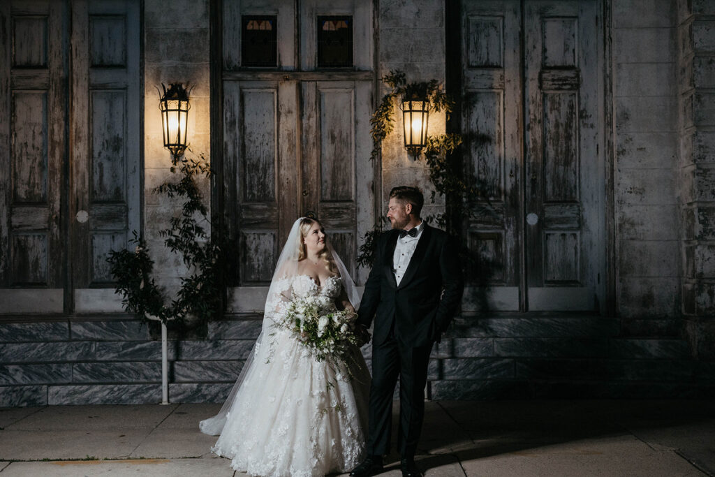 A newlywed couple holding hands and standing outside of a worn building 
