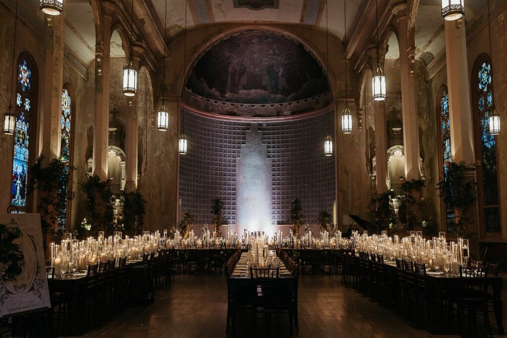 An elegant indoor wedding reception setup with long tables, covered in candles, set in a historic building with high ceilings and stained glass windows.
