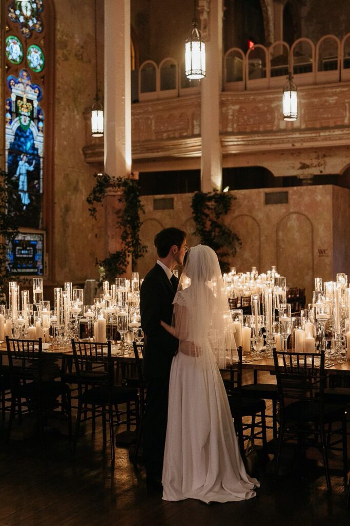 The bride and groom embracing among the candlelit tables in a historic, beautifully decorated reception hall.
