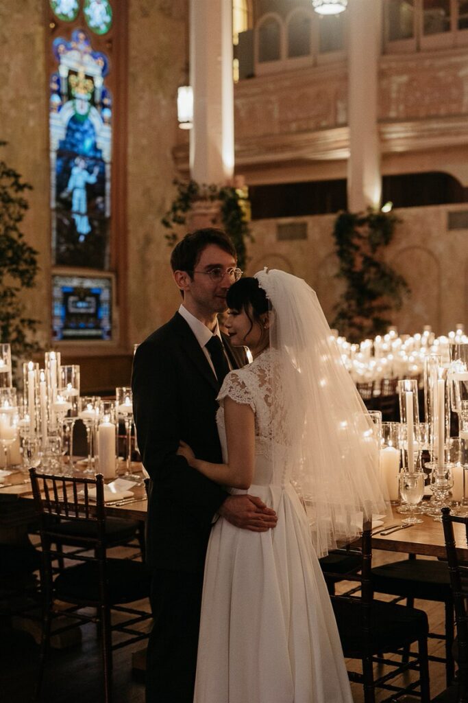 The bride and groom embracing among the candlelit tables in a historic venue, with stained glass windows in the background.
