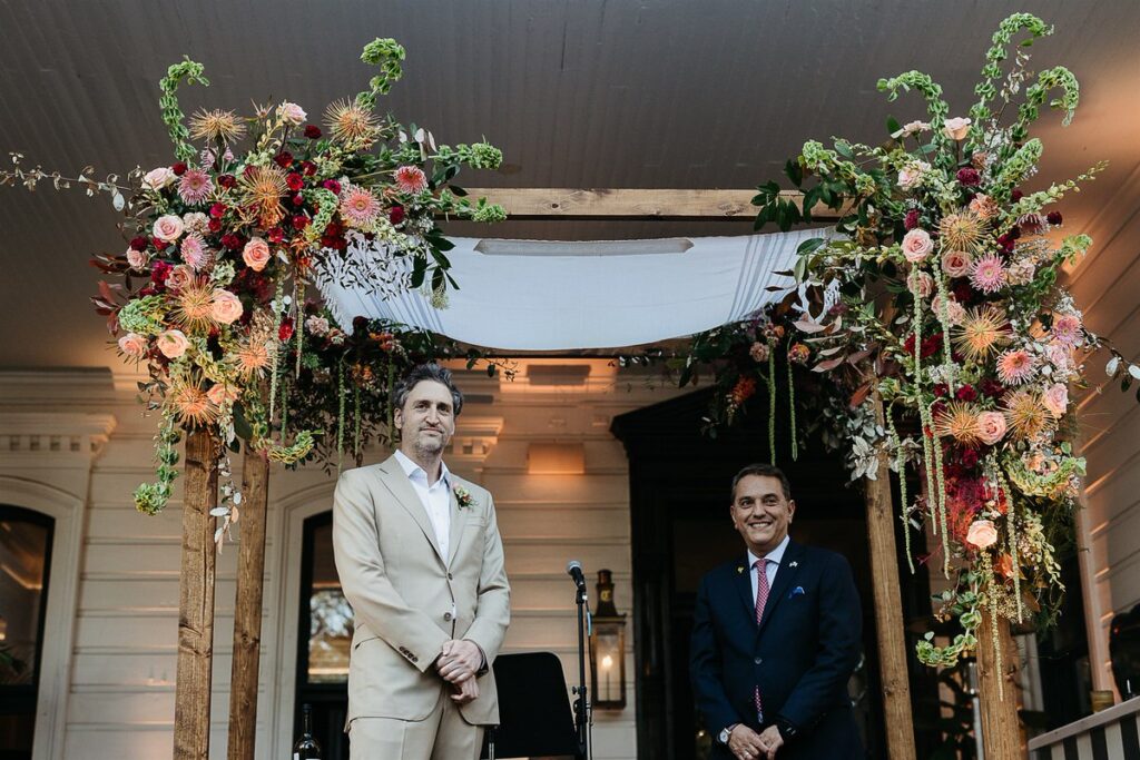 Two men, one in a beige suit and the other in a dark suit, stand under a floral-decorated wedding chuppah.