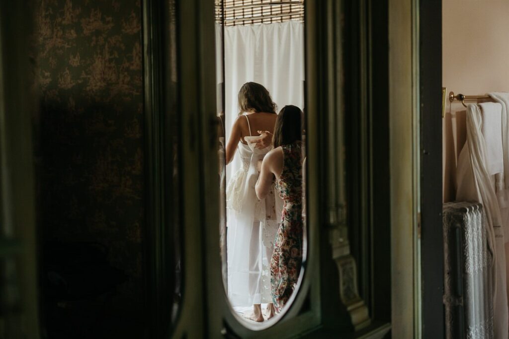 A bride in a white wedding dress is getting ready with the help of another woman, reflected in a mirror.
