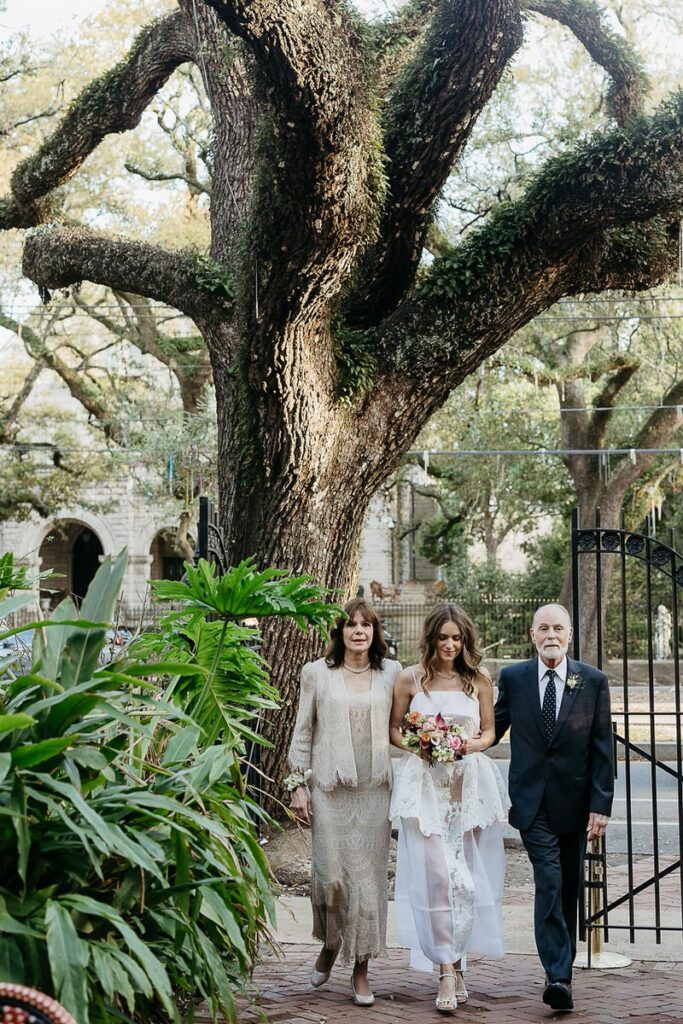 Bride walking down a garden path with a woman and an elderly man, surrounded by lush greenery and a large tree.
