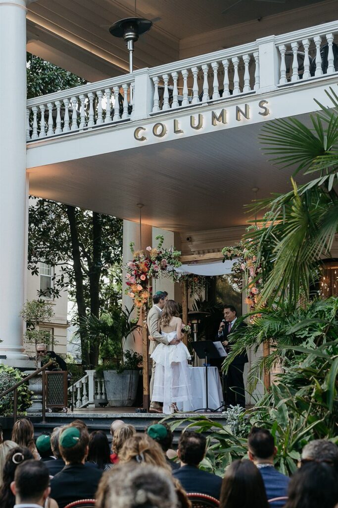 Wedding ceremony taking place on the porch of a building named "Columns," with the couple standing under a floral-decorated chuppah.
