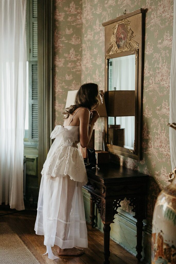 Bride in a white dress with a large bow on the back, standing in front of a mirror in a vintage room.
