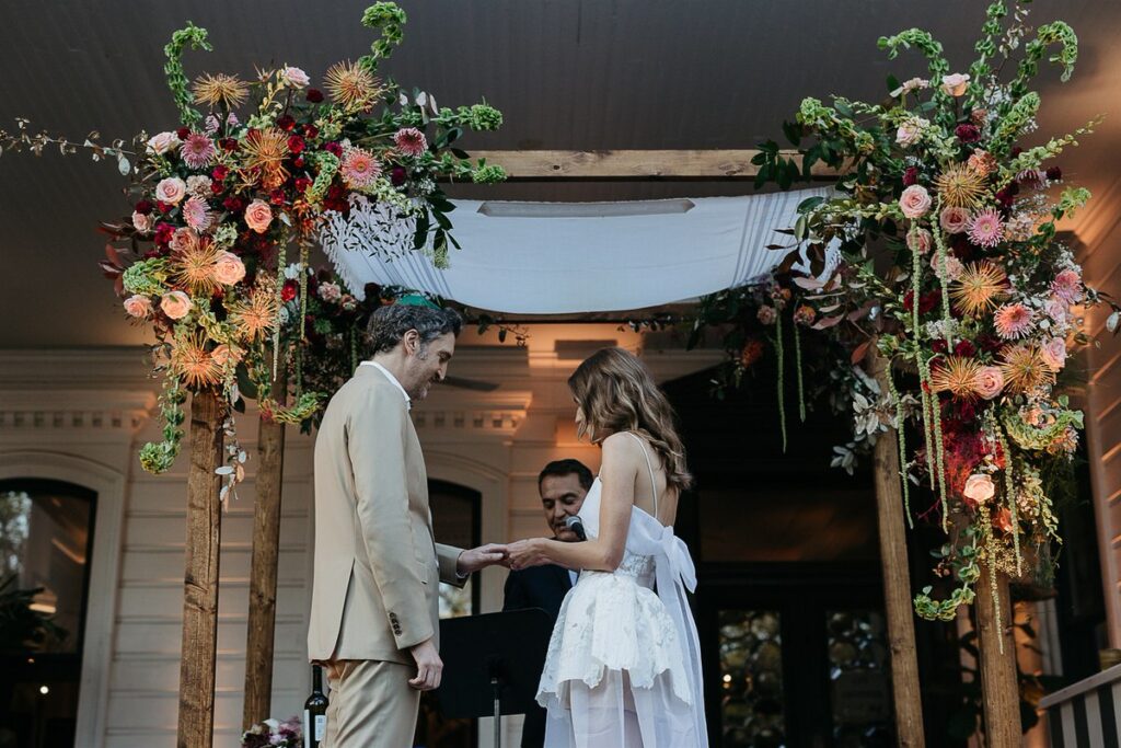 Bride and groom exchange rings under a chuppah adorned with vibrant floral arrangements.
