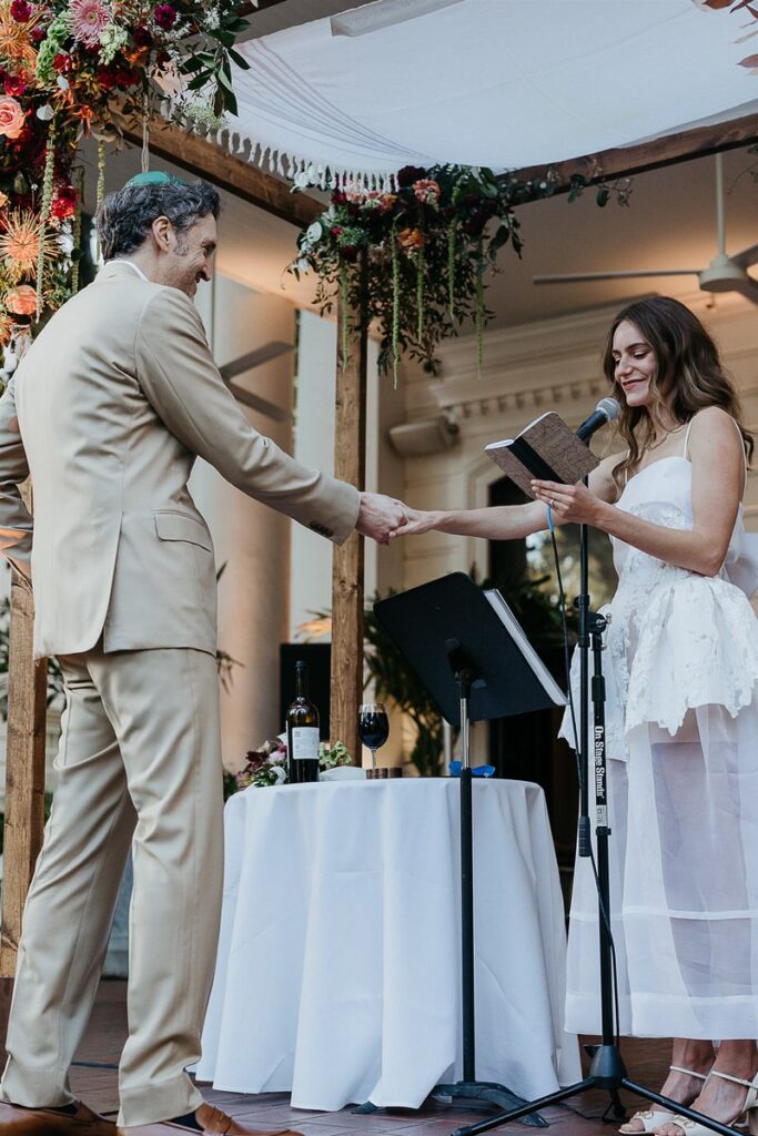 Groom holds the bride's hand while she reads from a book during their wedding ceremony.
