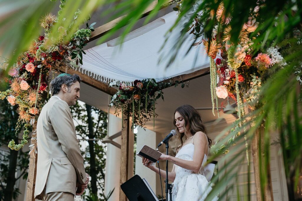 Bride reading vows with a microphone, standing under a chuppah with lush floral decor, the groom listening attentively.
