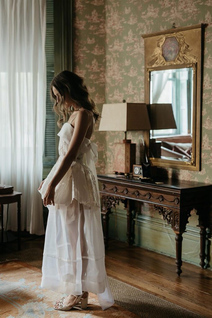 Bride standing in a vintage room, looking down, wearing a white dress with lace details and sheer fabric.
