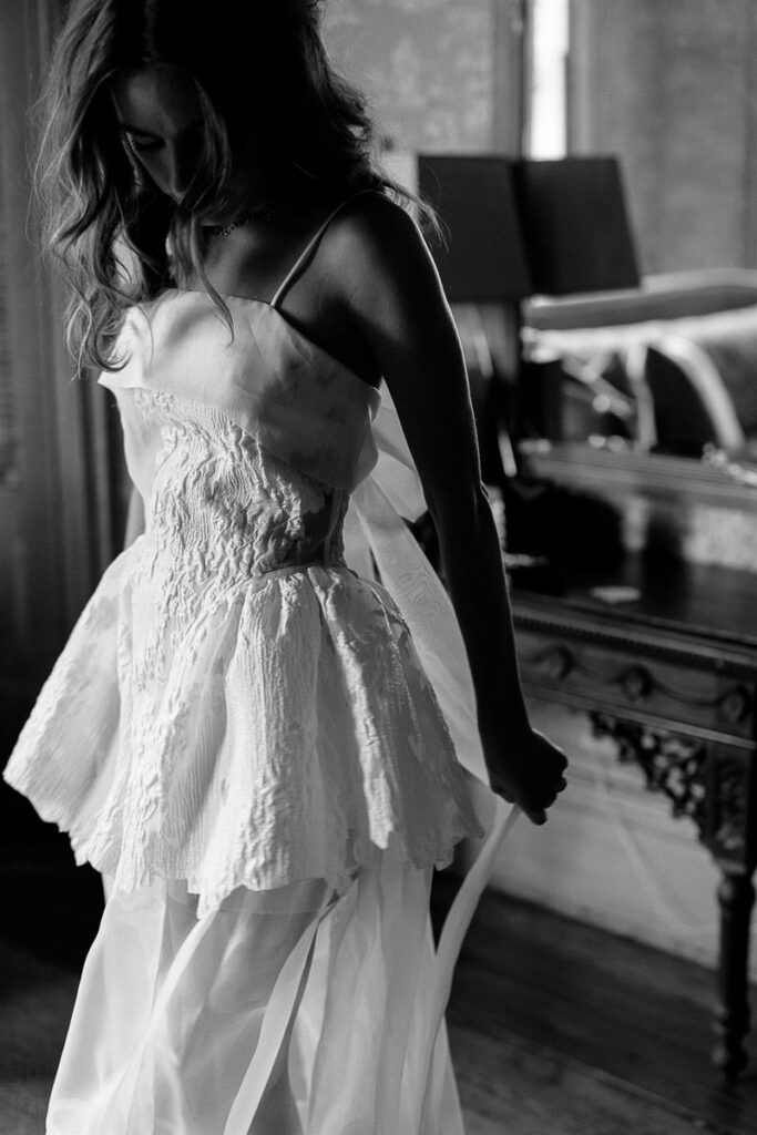 Black and white image of a bride in a detailed wedding dress, standing in front of a mirror.
