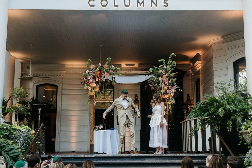 Wedding ceremony under the chuppah on the porch of a venue named "Columns," with guests watching.
