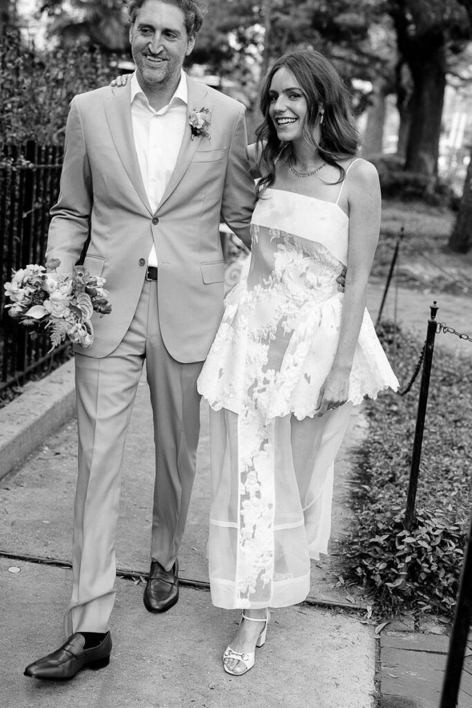 Black and white photo of the bride and groom walking together, smiling, with the groom holding a bouquet of flowers.
