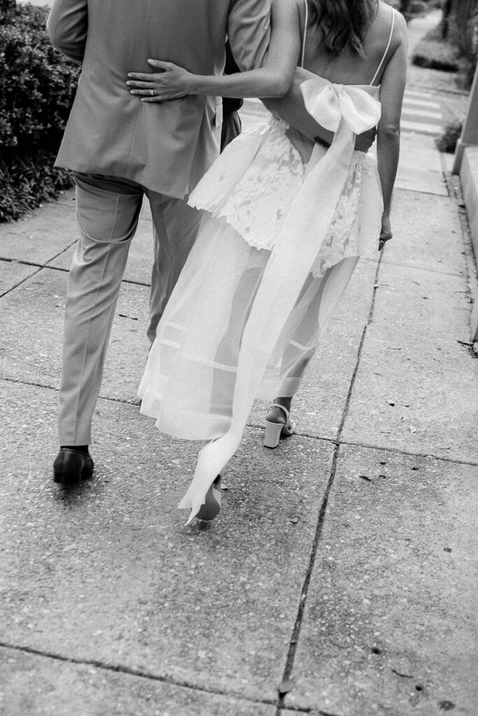 Back view of a couple walking on a sidewalk, with the bride's dress featuring a long white ribbon flowing behind her.
