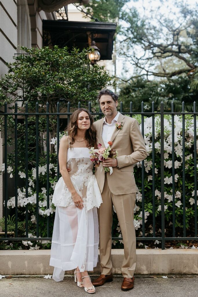 The bride and groom posing together outside, with white flowers and greenery behind them.
