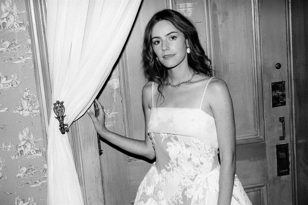 The bride standing inside by a doorway next to a white curtain looking at the camera.
