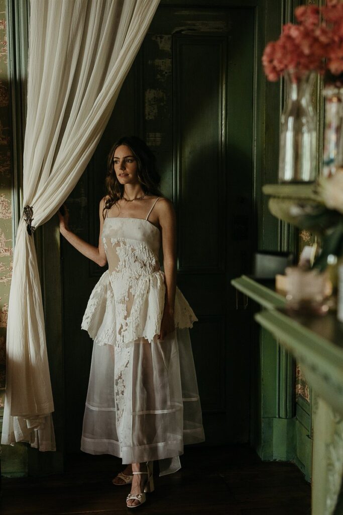 The bride standing inside next to a white curtain, looking into the distance.
