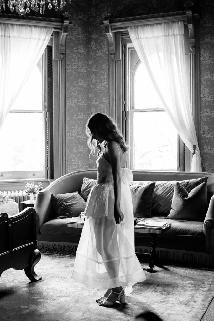 The bride in profile, standing in a room with a sofa and curtains, backlit by window light.
