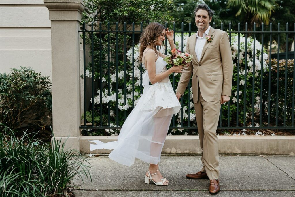 The bride and groom smiling and holding hands outside, with white flowers in the background, the bride's dress flowing in the wind.
