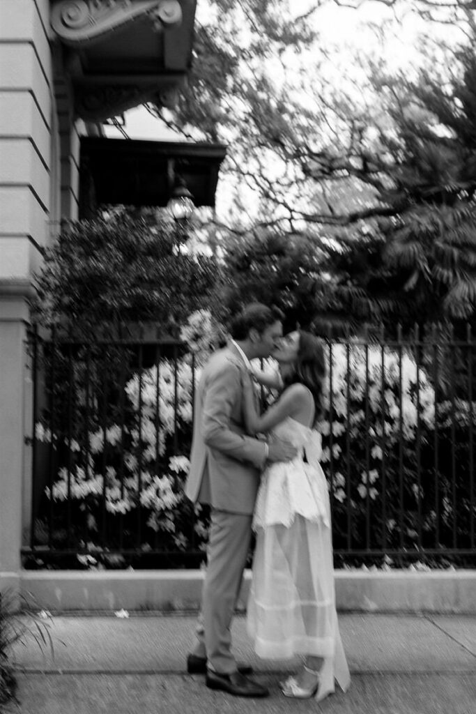 A slightly blurred photo of the bride and groom kissing outside, with greenery and flowers behind them. ​​