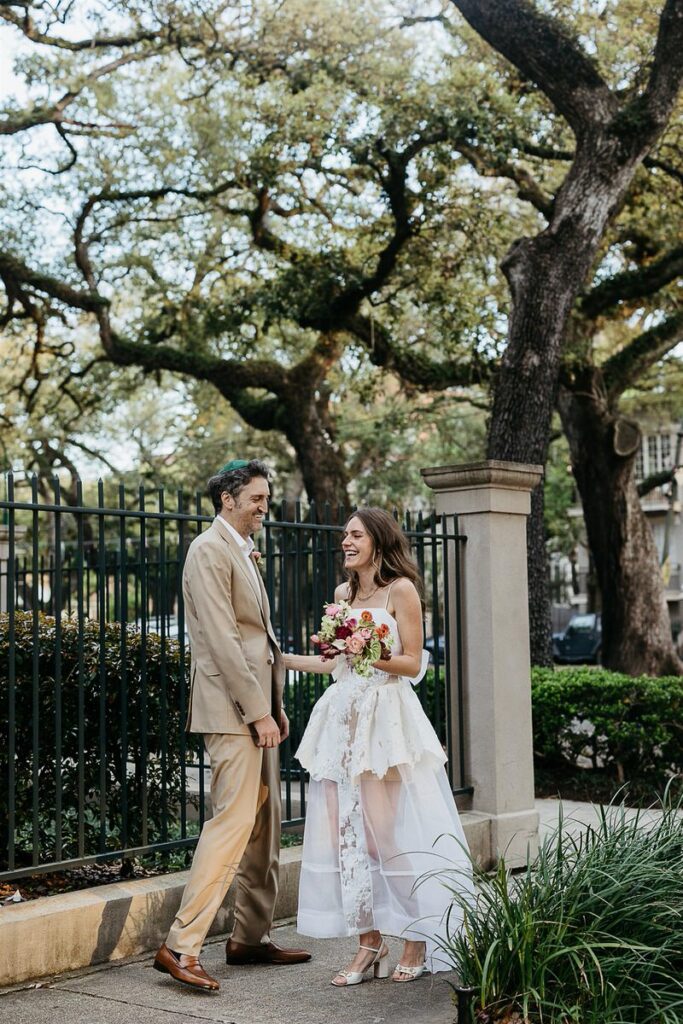 The bride and groom laugh together outside, surrounded by greenery and a black iron fence.





