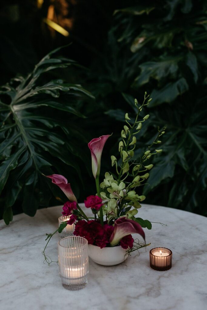 A small floral arrangement with candles on a marble table, set against a background of large green leaves.
