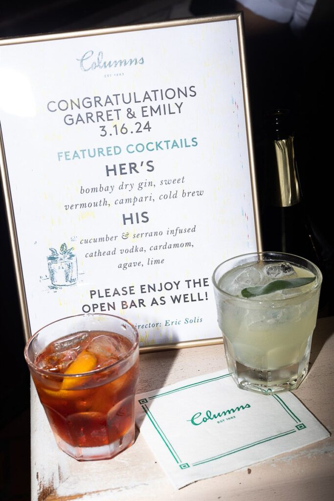 A sign at the bar featuring congratulations for Garrett and Emily, with their featured cocktails listed and drinks in the foreground.
