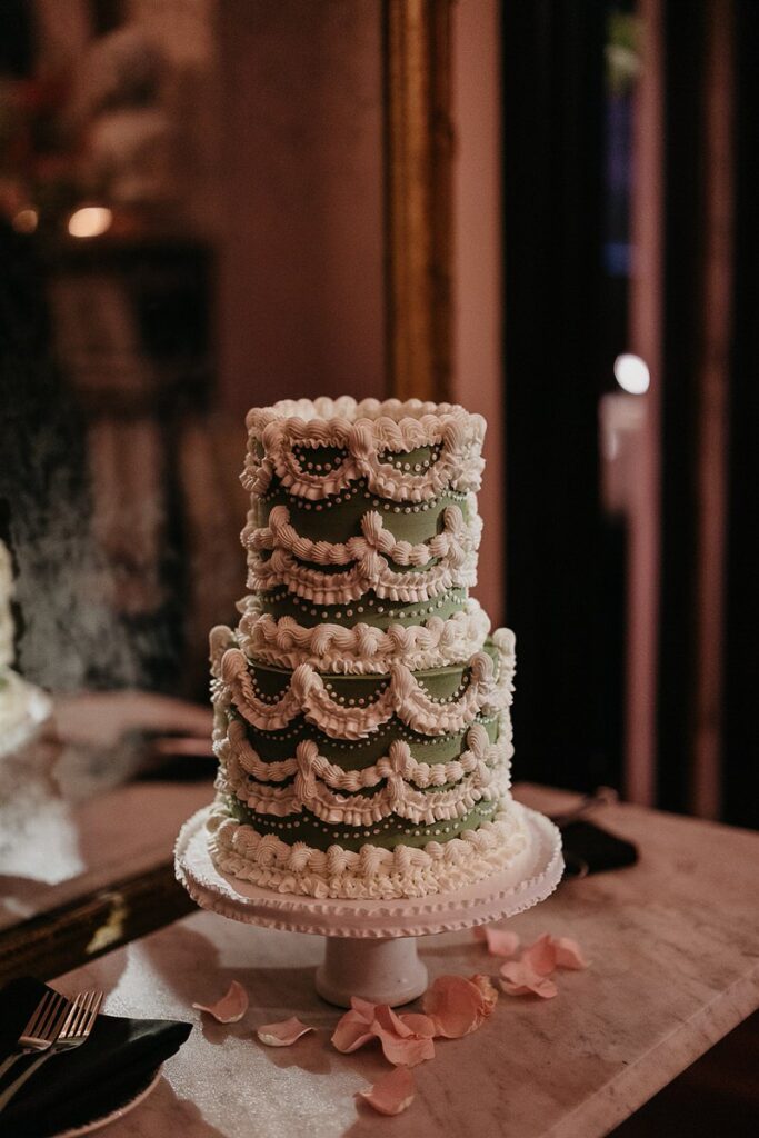 A three-tiered wedding cake with intricate white frosting decorations on a green background, sitting on a table with pink rose petals.
