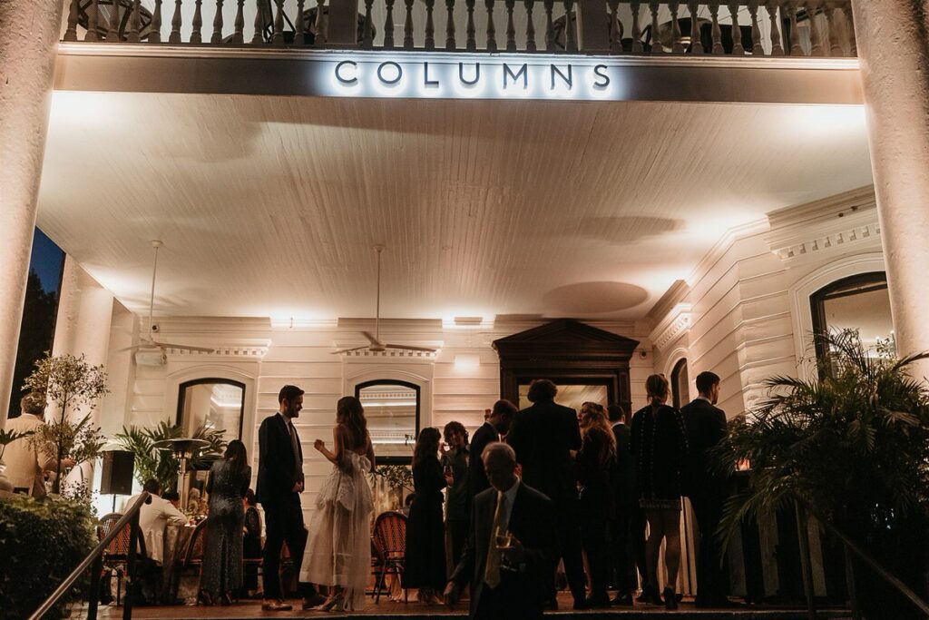 Evening view of the Columns venue with guests standing on the porch, the venue sign illuminated above.
