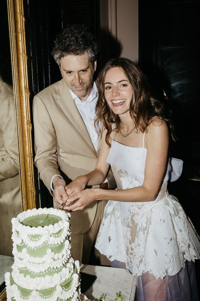 The bride and groom cut the wedding cake together, both smiling.
